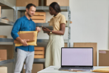 Obraz premium Blank laptop screen in warehouse with people working in background, small business and shipping service concept, copy space