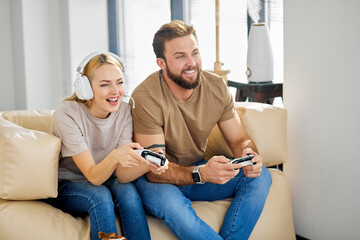 Cheerful man and woman sitting on sofa in living room playing computer video game, holding controller in hands, having fun, smiling. Side view portrait, copy space. Modern technologies