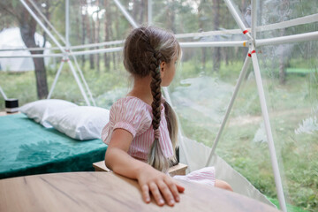 Cute girl looking through transparent bell tent with comfortable bed in forest, glamping hotel, luxury travel, glamourous camping with amenities, dome tent, feel at home in great outdoors lifestyle