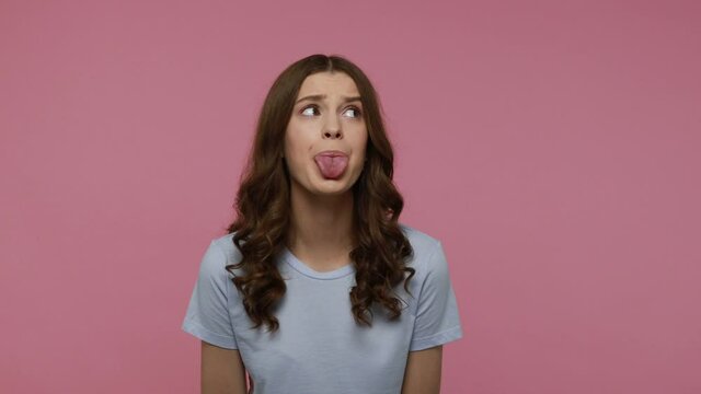 Carefree lovely girl with wavy hair having fun in front of camera, showing tongue out, behaving naughty disobedient with childish mood.Indoor studio shot isolated over pink background.