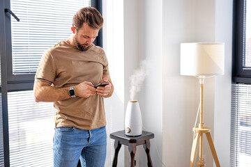 Smart air humidifier emitting water vapor on table, While man use smartphone, living in smart...