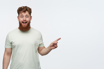 indoor shot of young bearded redhead guy, wears blank t-shirt, pointing with a finger at right side copy space, smiling and has amazed facial expression. isolated over white background.