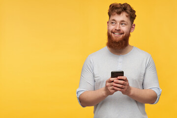 young redhead male with a big beard smiles and holds smartphone in his hands. looking aside to the right at copy space. isolated over yellow background