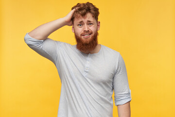 indoor shot of young bearded guy with red hair, wears blank t-shirt, looking irritated into camera , touching his head. isolated over yellow background