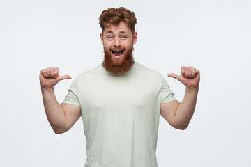 portrait of positive cheerful male with big beard and red hair wears blank t-shirt, pointing with thumbs himself, smiles broadly and has happy facial expression. isolated over white background
