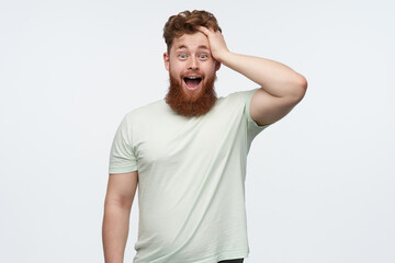 young amazed redhead bearded man, keeps his mouth and eyes open widely, hold his head with hand. isolated over white studio wall.