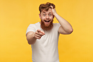 Fototapeta na wymiar portrait of cheerful positive man with red beard wears blank t-shirt, laughing and point with a finger at the camera. isolated over yellow background