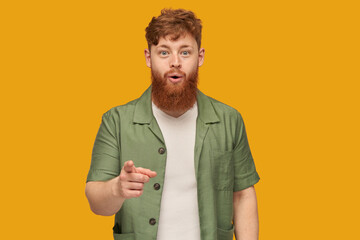 Indoor shot of young handsome redhead man with big beard, looking directly into camera with shocked, astonished facial expression, pointing with a finger at you. Isolated over yellow background.