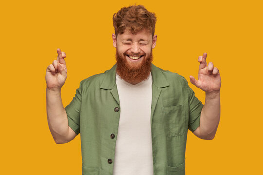 indoor shot of young bearded male, with ginger hair, crossed his fingers in praying position, keeps his eyes closed while waiting exams result. Isolated over yellow background.