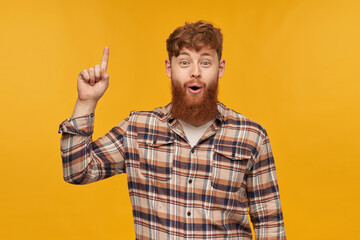 Fototapeta premium Young positive male, with big red bear, looks into camera with shocked facial expression, point with a finger upwards and have new idea for start-up. Isolated over yellow background