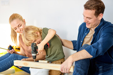 boy kid sit on floor and help parents assembling wooden board with power drill tool. Young mother...