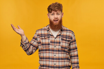 Indoor shot of sad depressed young male, looks into camera with confused facial expression, raised his hand, isolated over yellow background