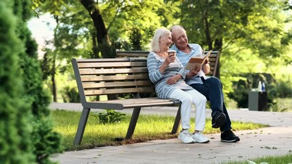 A happy elderly couple man and woman are sitting on the bench while reading book in the park - Powered by Adobe