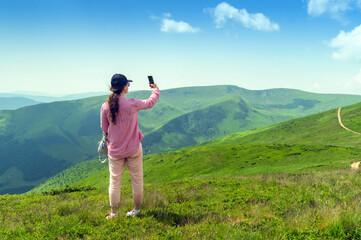 Fototapeta na wymiar Girl tourist is relaxing in nature and uses the Internet on a smartphone. The girl traveler photographs a panorama of the mountains on a mobile phone. Mountain beautiful landscape