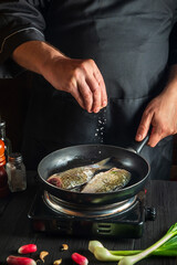 Professional chef prepares fresh fish in pan sprinkling salt with the ingredients. Preparing to cook fish food. Working environment in the restaurant kitchen.