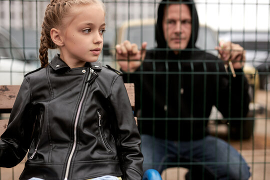 Maniac man watches through fence at little girl sitting on playground alone, child abuse concept. Caucasian pedophile guy in black wear want to commit crime, look at girl with interest, focus on girl