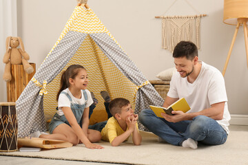 Father reading book to children near toy wigwam at home © New Africa
