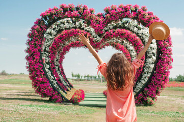 young woman in pink dress and hat stands and rise her hands up on background of flower heart arches...