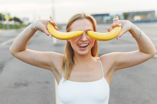 Frivolously and mischievous young woman with braces on teeth holding bananas on her face has fun outdoors. Emotions
