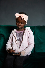 Low-key portrait of beautiful black South African girl wearing pink outfit with a pink bow on her head on a dark charcoal background, using phone while sitting on emerald green couch.