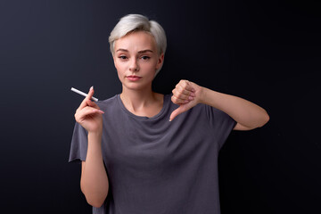 Obraz premium woman show dislike gesture, refuse to smoke cigarettes. Unhealthy lifestyle concept. short-haired caucasian lady in casual t-shirt look at camera seriously, say no smoking. Isolated black background