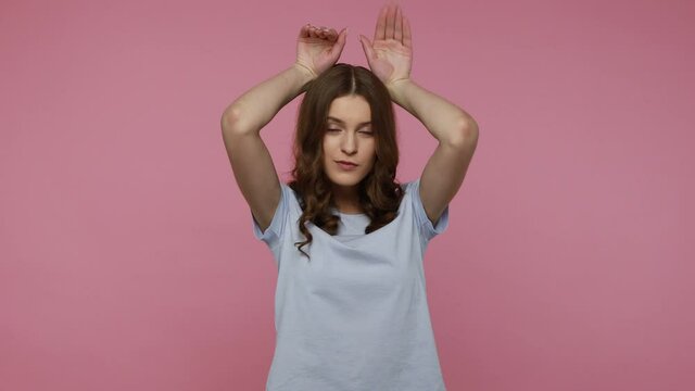 Funny adorable teenager with long black hair in T-shirt showing bunny ears with arms, rabbit gesture and having fun, looking cute childish. Indoor studio shot isolated over pink background.