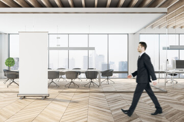 Businessman in suit walking in wooden coworking meeting room interior with empty mock up poster, daylight, furniture and equipment. Design and ceo worker concept.