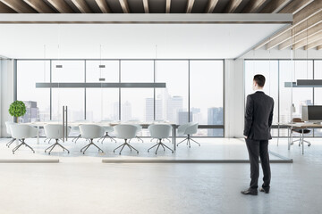 Thoughtful young businessman in suit standing in concrete coworking meeting room interior with city...