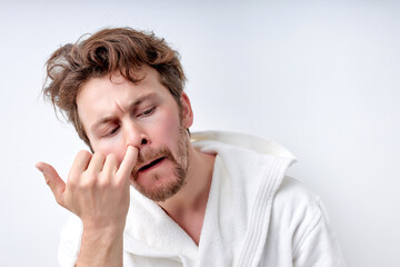 crazy handsome bearded young man in bathrobe drilling nose with funny face. caucasian male having bad habit. indoor studio shot, isolated on white background. copy space