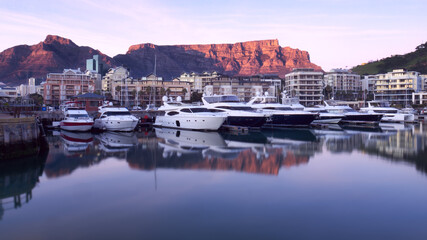 Obraz premium Table Mountain sunrise is reflected in the still waters of a marina for luxury motor yachts in Cape Town, South Africa