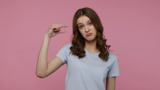 Pretty teenager girl in casual T- shirt showing small size, looking with disappointed expression, meaning little size, low rating, measuring scale. Indoor studio shot isolated over pink background.