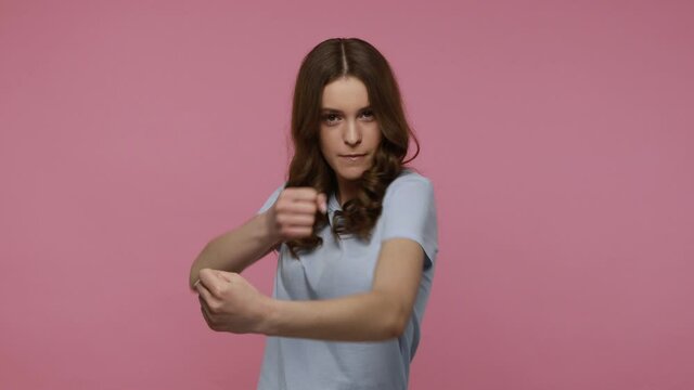 Let's fight! Young woman with dark wavy hair in casual T-shirt looking at camera, ready to boxing, threatening to punch, fighting spirit, Indoor studio shot isolated over pink background.