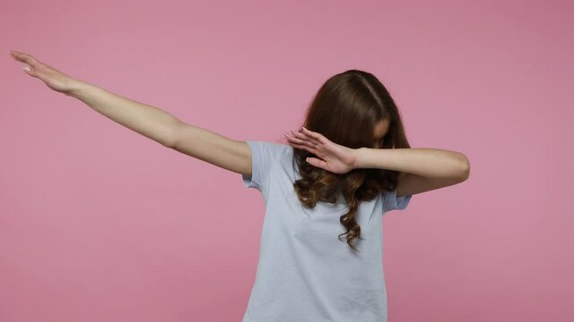 Satisfied joyful teenager girl in T-shirt celebrating success with dab dance move, famous internet meme of triumph, performing dabbing trends. Indoor studio shot isolated over pink background.