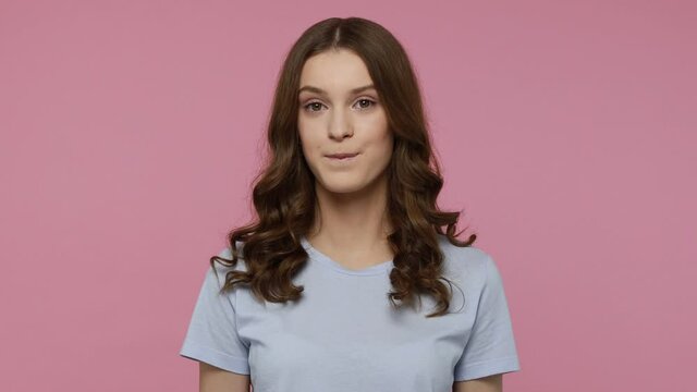 Portrait of beautiful teenager girl wearing blue T-shirt winking and looking at camera, flirting expression, cheering up with optimistic positive mood. Indoor studio shot isolated over pink background