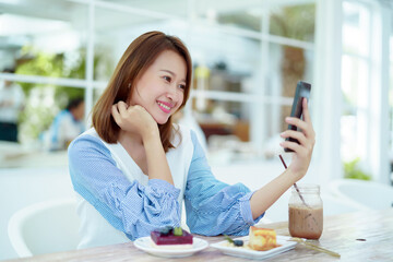 Portrait A beautiful Asian woman in a white shirt sits happily using her cell phone to take a selfie in a bakery.