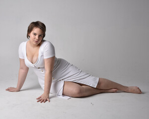 Full length portrait of young plus sized woman with short brunette hair,  wearing l tight white body con dress,  kneeling pose with gestural hands on ground with light studio background.