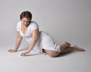 Fototapeta premium Full length portrait of young plus sized woman with short brunette hair, wearing l tight white body con dress, kneeling pose with gestural hands on ground with light studio background.