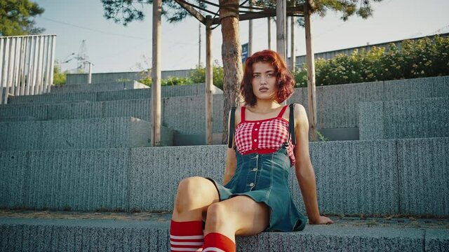 Young beautiful redhead pin up woman sitting in urban park, posing like fashion model, tracking shot