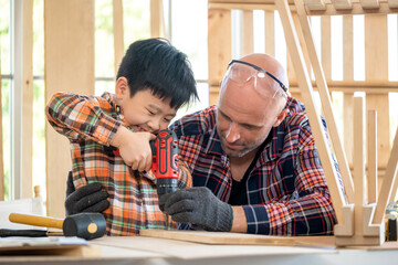 Senior carpenter wearing glove and teach a young boy to use screwdriver in the modern wood workplace classroom. Man support kid and concentrate on work. Education and learning concept. DIY handcraft
