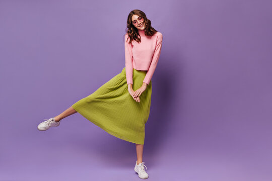 Pretty curly woman rises leg. Attractive young girl in green midi skirt and pink sweater smiles and moves on purple background.