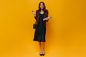 Woman with wavy hair holds books on orange background. Attractive lady in silk dress and checkered jacket smiles on isolated.