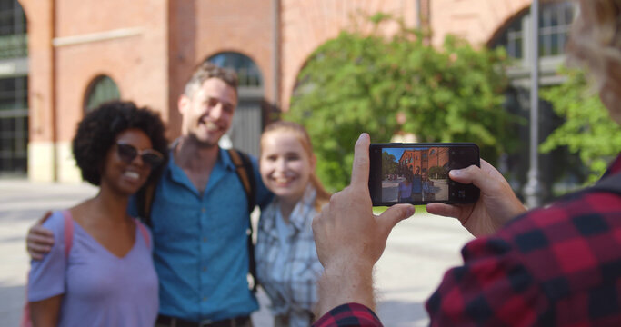 Over shoulder view of young man taking photo of happy friends on smartphone traveling together