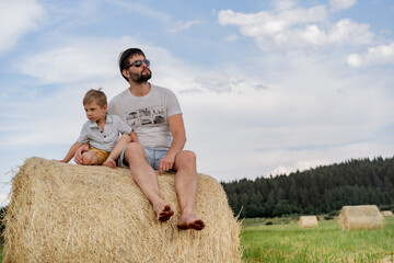 Naklejka na ściany i meble portrait of man and his little son sitting on round haystack in a green field on sunny summer day.