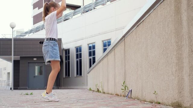 Schoolgirl blogger dances. Blonde teenager with long hair in ponytail performs squat dance jumping moves for blog using phone put on paved street path by concrete fence