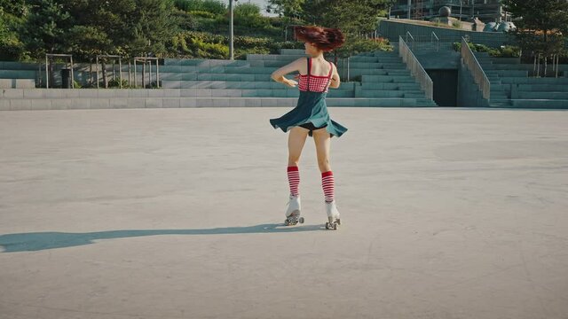 Young gorgeous woman wearing bright pin up dress rinking on roller skates in urban park, dancing and moving gracefully