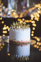 Birthday cake with candles, garland with bright bokeh lights on the background. The white cake is decorated with colored sprinkles and stands on a reflective surface. © Evgeniia Biriukova