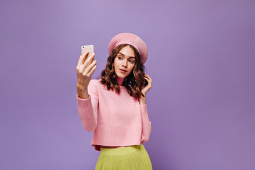 Obraz na płótnie Canvas Elegant woman holds phone and takes selfie. Young curly Parisian lady in pink beret, sweater and green skirt poses on purple background.