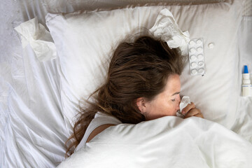 A sick young woman in bed, lying with her head under the blankets and a pile of tissues next to her...