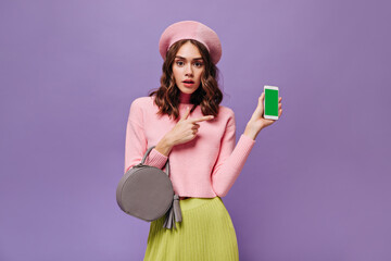 Brown eyed woman points at phone screen. Happy lady in pink beret, sweater and green skirt holds grey handbag on purple background.