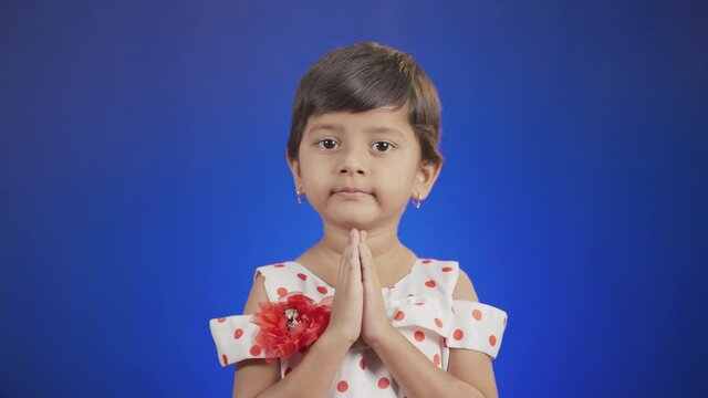 young girl kid on blue background doing namaste by looking camera - Concept of respectful greetings or saying hi or hello in indin culture